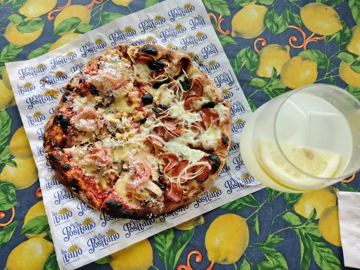 Pizza sobre o papel da Villa Positano, numa mesa com toalha de limões, ao lado de um spritz