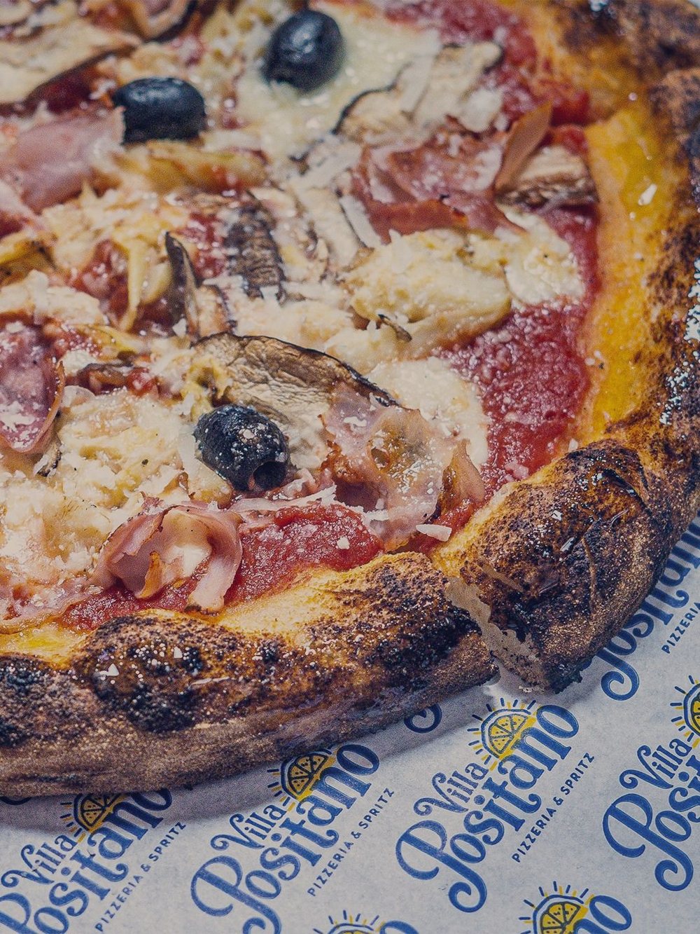 Pizza com presunto, azeitonas e cogumelos, servida sobre o papel da Villa Positano